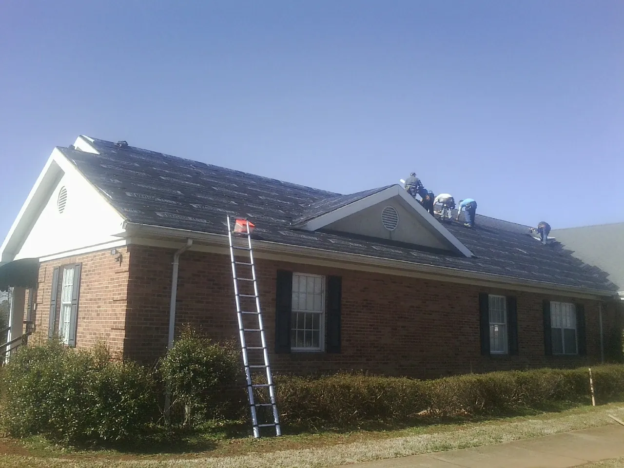 Crew tearing shingles off a brick ranch roof with a ladder set — Metal Roof Repair work by our crew in Eldred