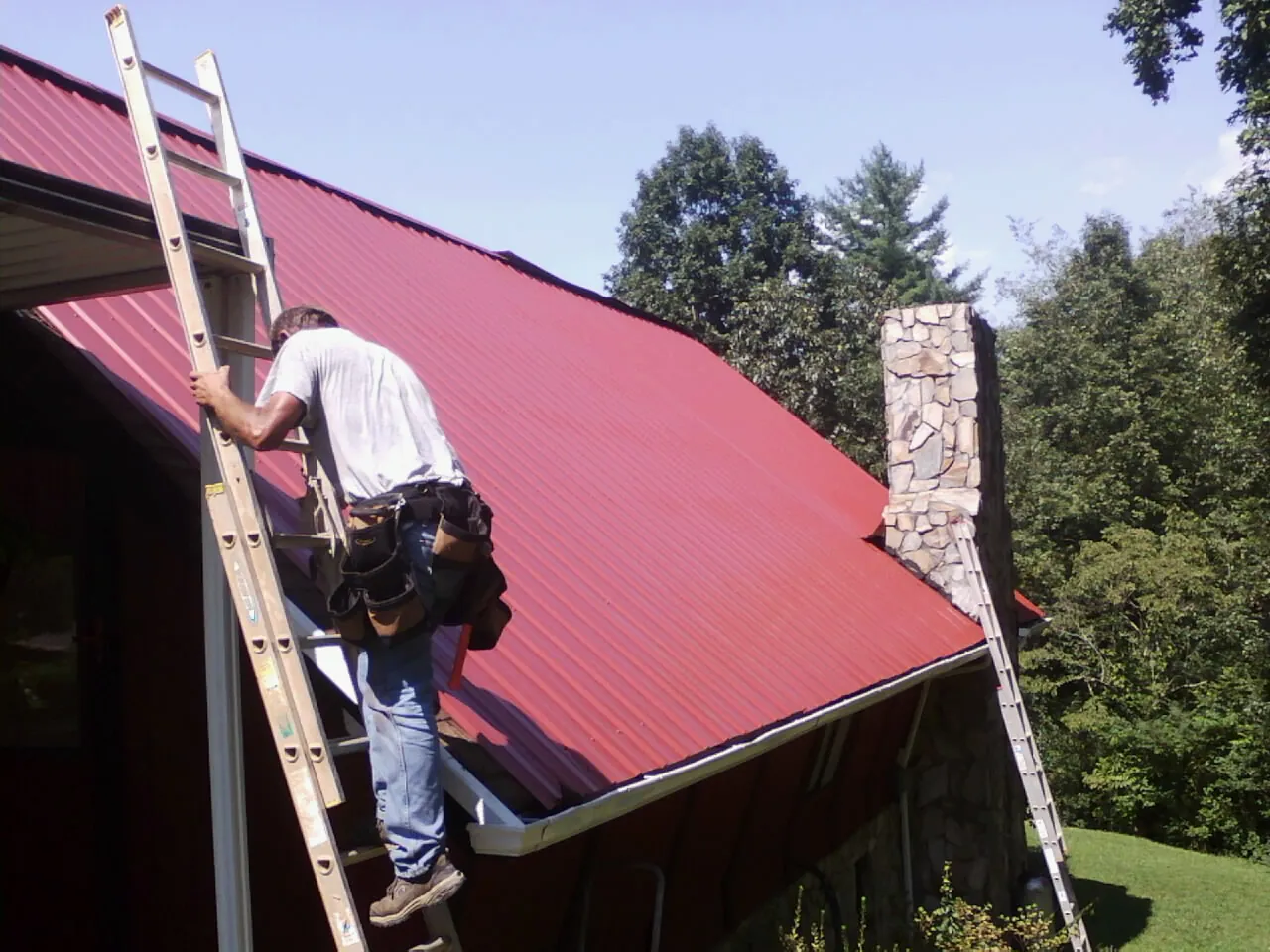 Roofer on a ladder beside a red metal roof and stone chimney — Soffit & Fascia Repair by Roofing Brothers in Eldred